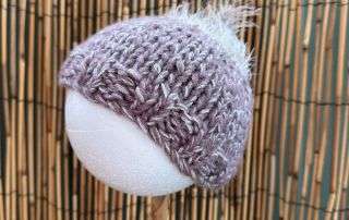 A lilac baby beanie handmade with a fluffy white tuft on top, sized for the tiniest of heads.