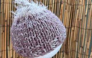 A lilac baby beanie handmade with a fluffy white tuft on top, sized for the tiniest of heads.
