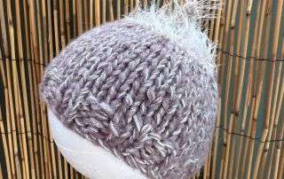 A lilac baby beanie handmade with a fluffy white tuft on top, sized for the tiniest of heads.