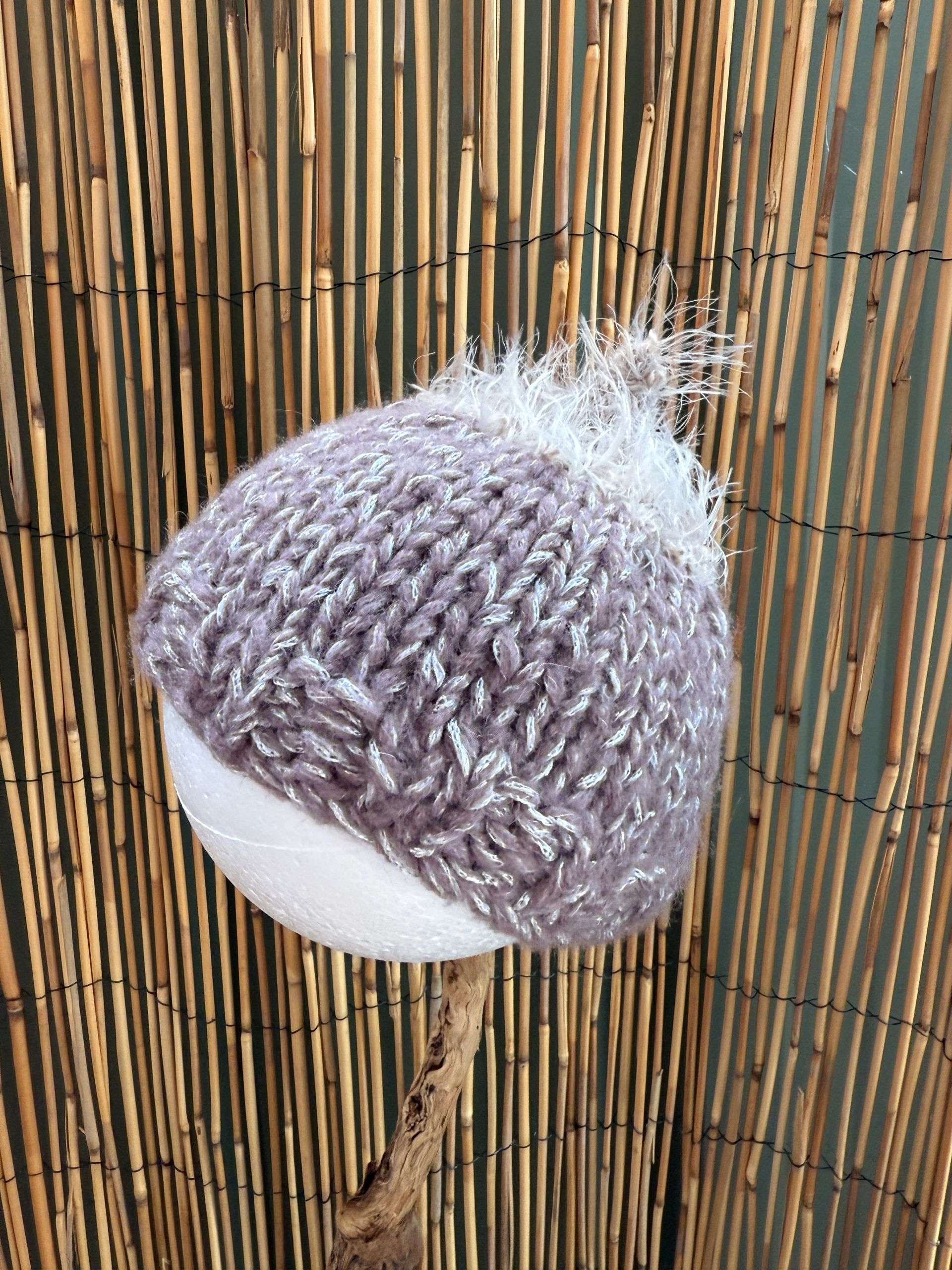 A lilac baby beanie handmade with a fluffy white tuft on top, sized for the tiniest of heads.