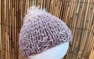 A lilac baby beanie handmade with a fluffy white tuft on top, sized for the tiniest of heads.